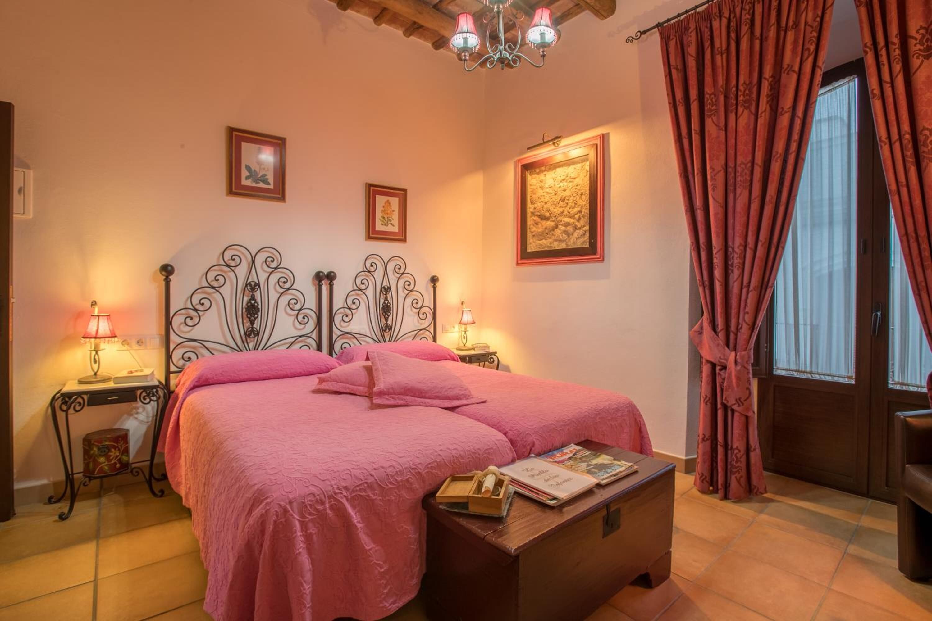 A bedroom with a bed, bedside tables, a nightstand, a wooden bench, a chandelier, and a window with curtains.