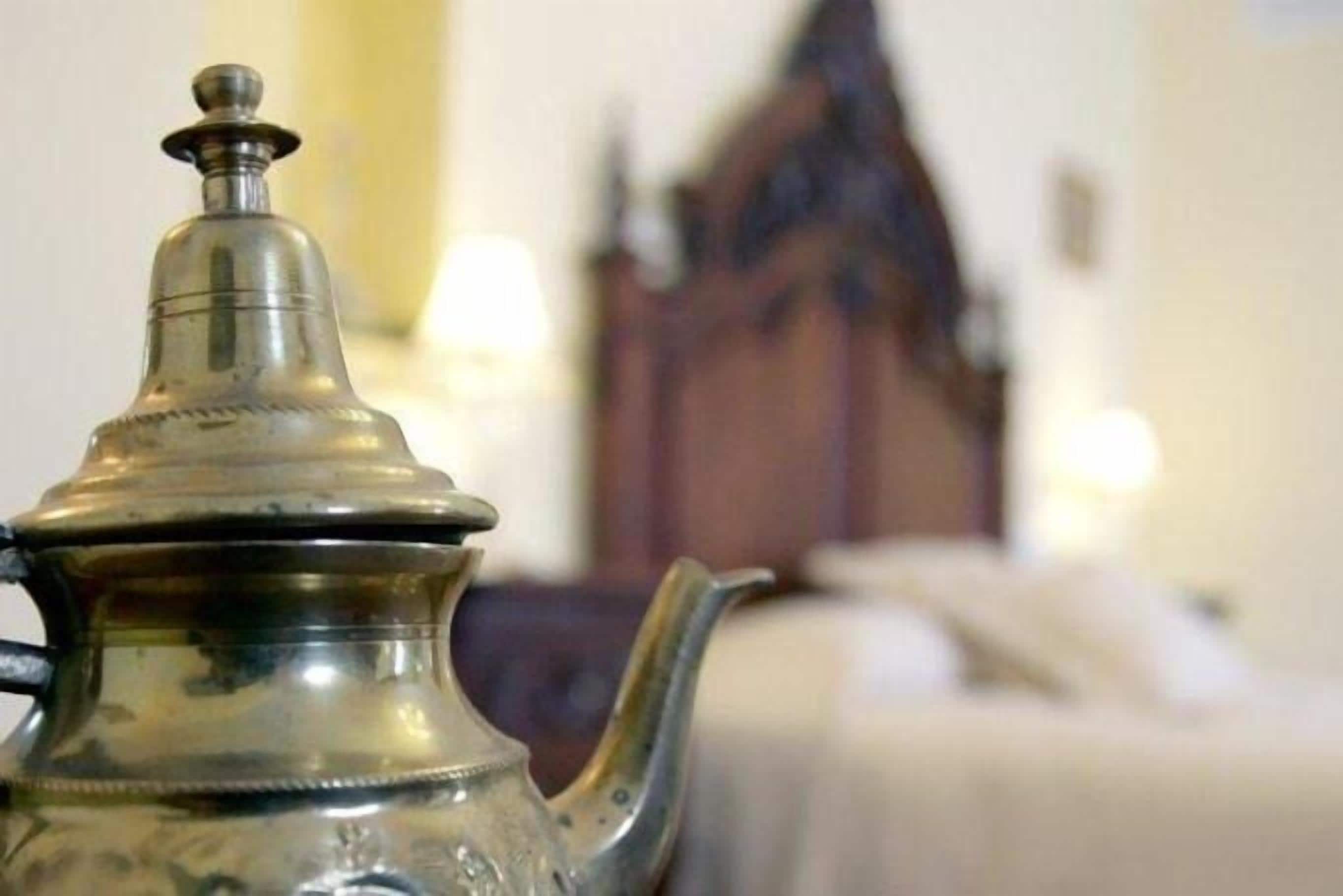Primer plano de una tetera de bronce con tapa y asa, sobre una superficie con fondo desenfocado que muestra un armario de madera y una cama.