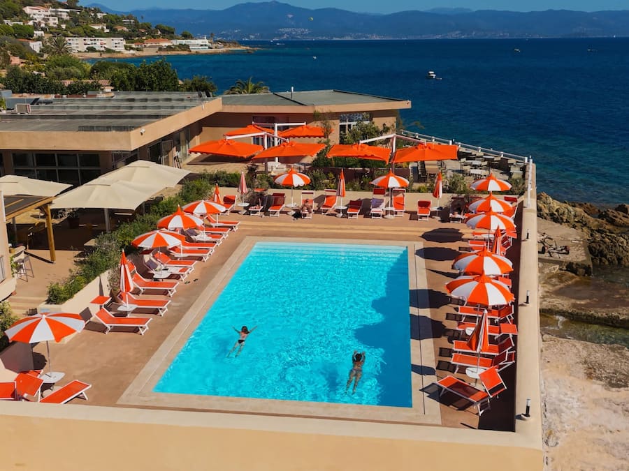 Une piscine dotée de chaises longues et de parasols, entourée de bâtiments et offrant une vue sur la mer.