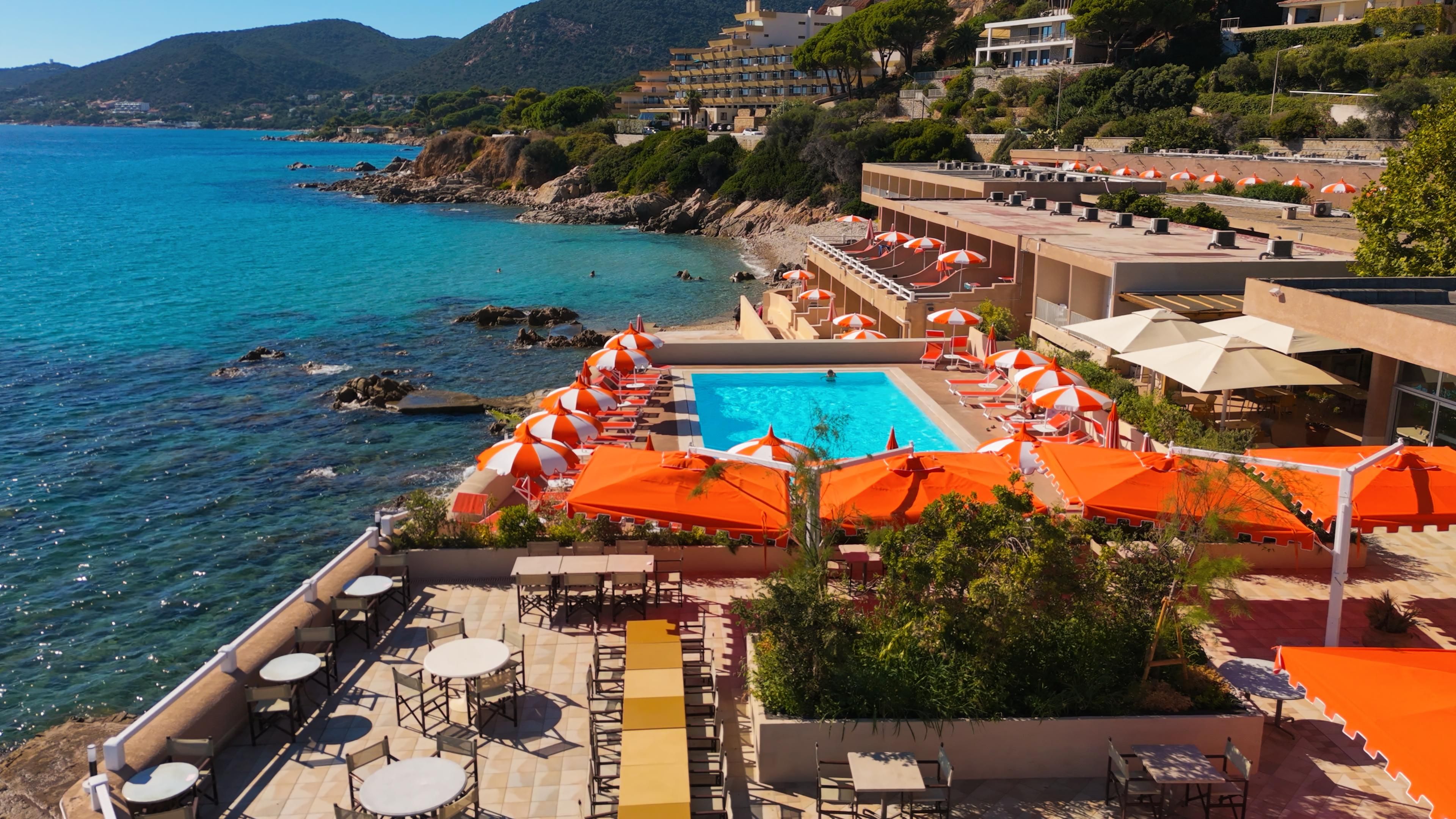 Ein Resort mit Swimmingpool, umgeben von Liegestühlen und Sonnenschirmen, mit Blick auf eine Küstenlandschaft mit klarem, blauem Wasser und Hügeln im Hintergrund.