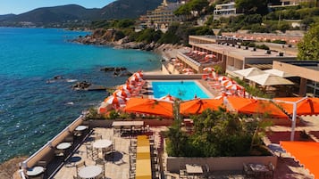 A resort with a swimming pool surrounded by sun loungers and umbrellas, overlooking a coastal area with clear blue water and hills in the background.