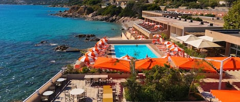 A resort with a swimming pool surrounded by sun loungers and umbrellas, overlooking a coastal area with clear blue water and hills in the background.