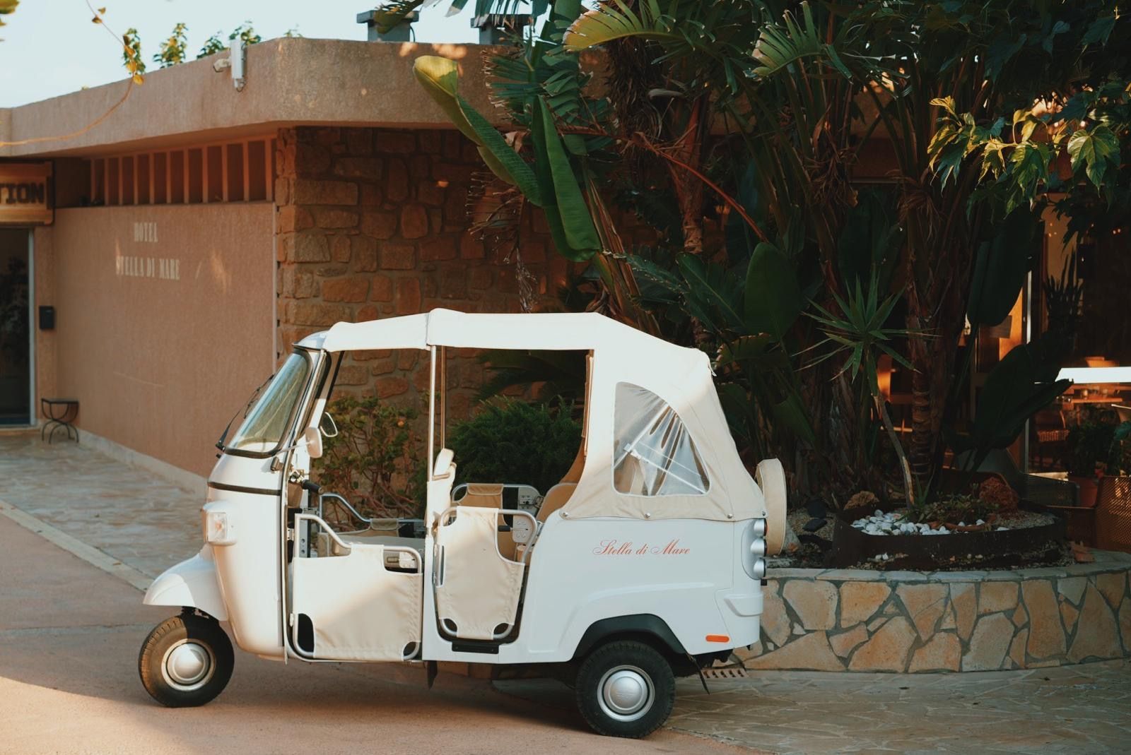 Un carrito de golf blanco estacionado frente a un edificio de fachada de piedra y plantas tropicales.