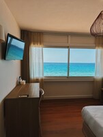 Habitación de hotel con un escritorio de madera, televisor montado en la pared, cama y un amplio ventanal con vista al mar.