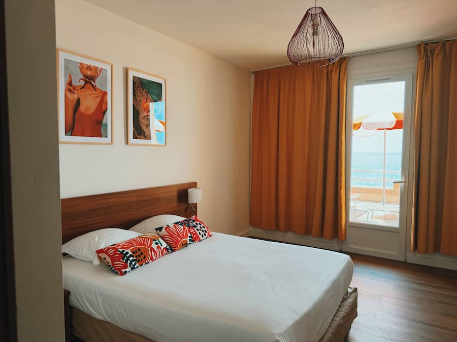 Une chambre d’hôtel avec un lit, deux tableaux encadrés au mur, un luminaire suspendu et une vue sur la plage par la fenêtre.