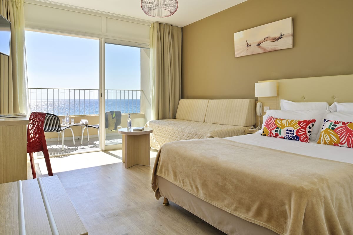 Une chambre d’hôtel avec un grand lit, un bureau, une chaise, une petite table et une vue sur la mer depuis les portes du balcon.