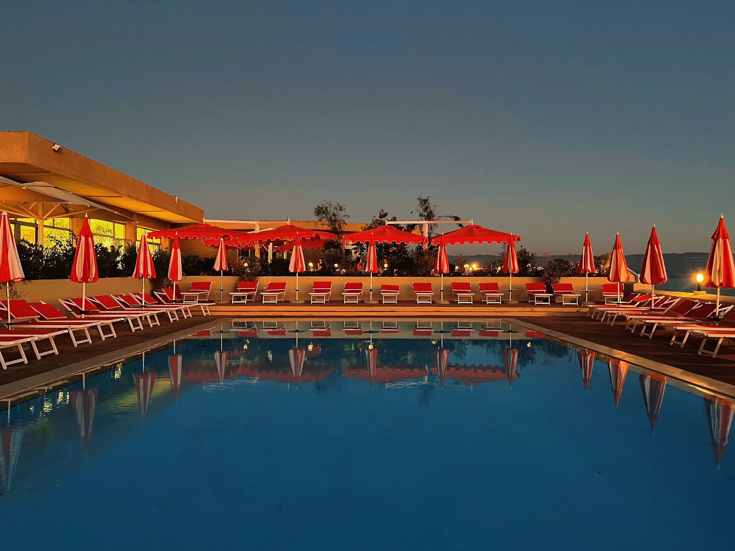 Una piscina con sombrillas rojas y sillones de descanso, rodeada por un edificio de techo plano.
