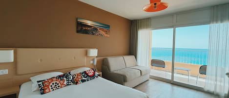 A hotel room with a bed, a sofa, a balcony with a view of the sea, and a painting on the wall.