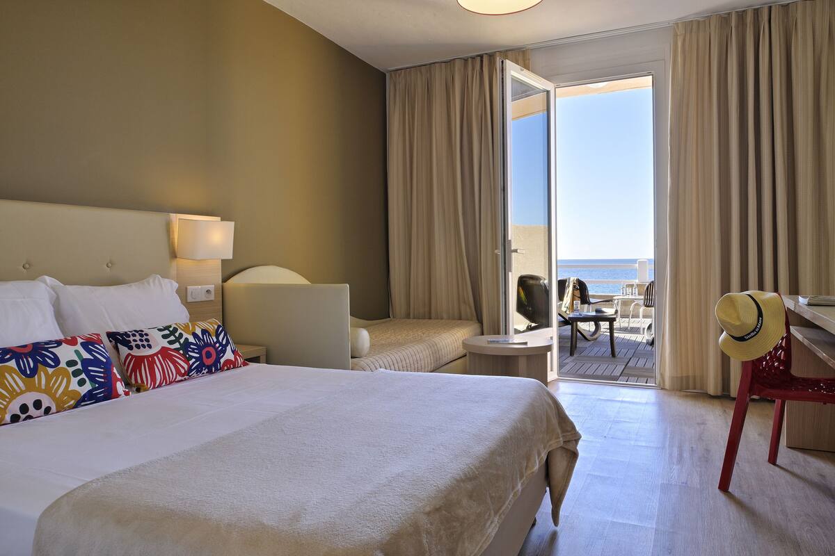 Une chambre d’hôtel avec un lit, un bureau, une chaise et une vue sur la mer, visible par une porte ouverte.