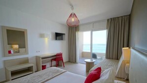 A hotel room with two beds, a desk, a chair, a TV, and a balcony with a view of the sea.