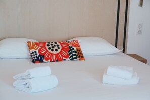 A neatly made bed with white linens, two pillows with a colorful floral pattern, and two rolled towels on the bed.