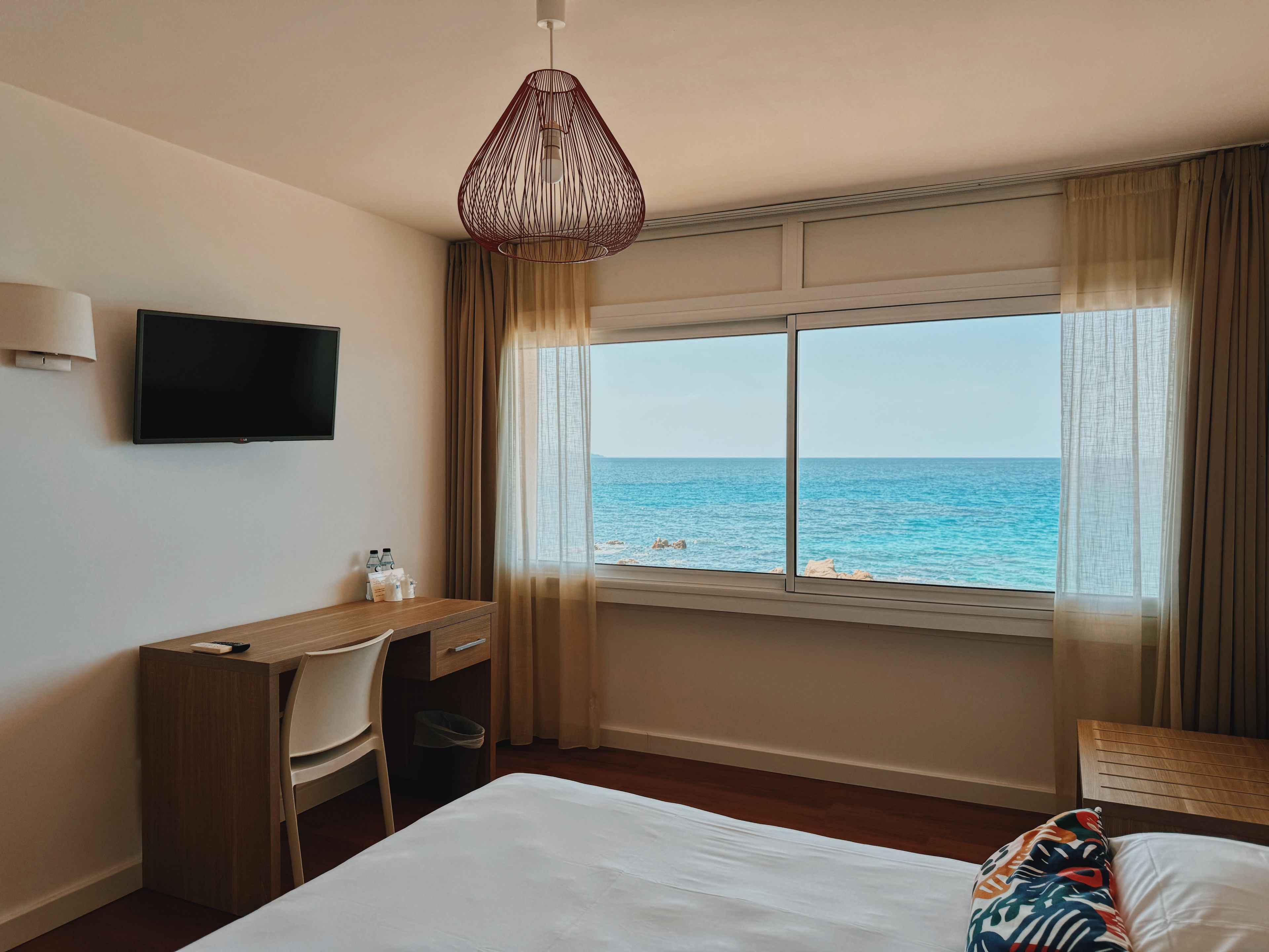 Ein Hotelzimmer mit Bett, Schreibtisch, Sessel, Fernseher und einem Fenster mit Meerblick.