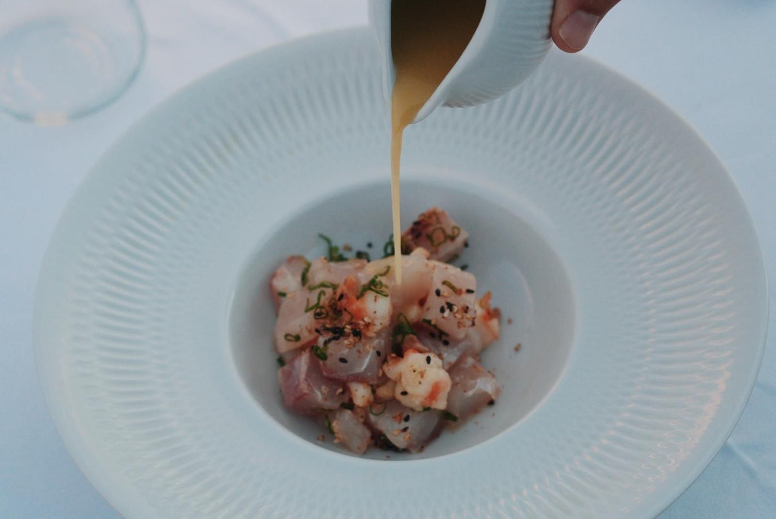 Un plato blanco con un plato de mariscos adornado con hierbas y especias, al que se le rocía una salsa.