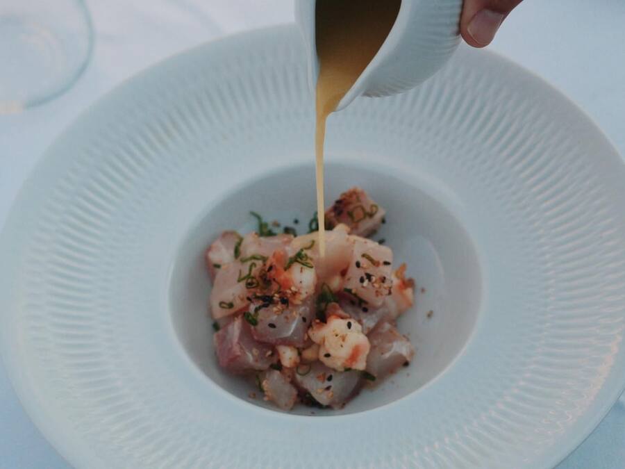 A white plate with a seafood dish garnished with herbs and spices, being drizzled with a sauce.