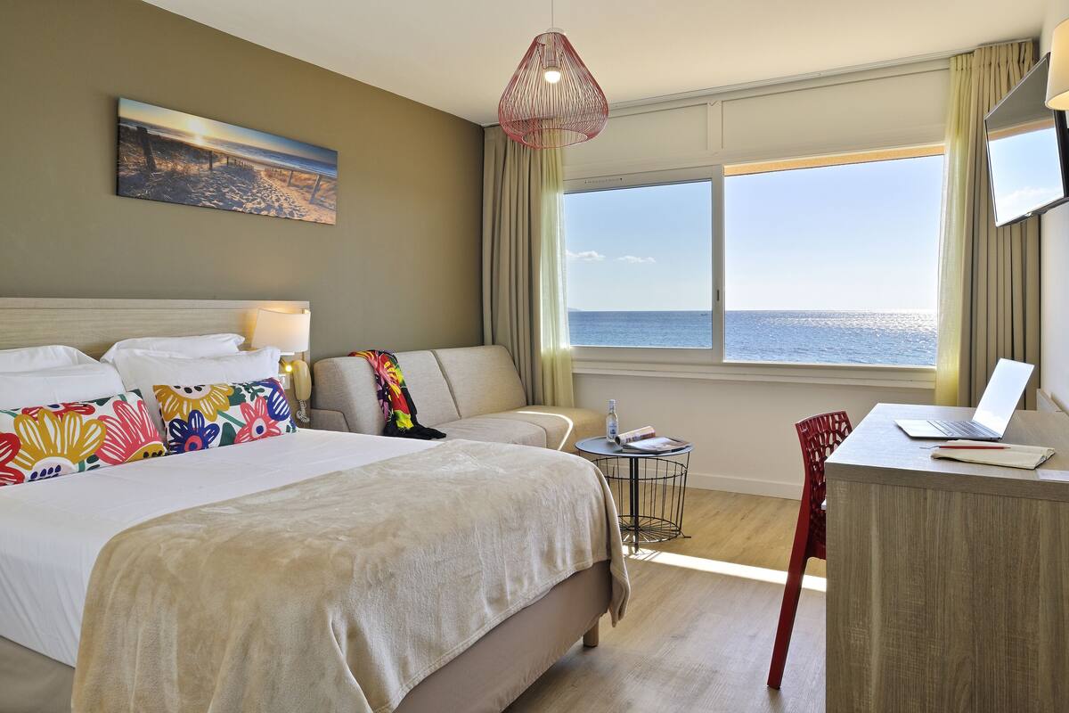 Une chambre d’hôtel avec un lit, un bureau, un ordinateur portable et une vue sur la mer.