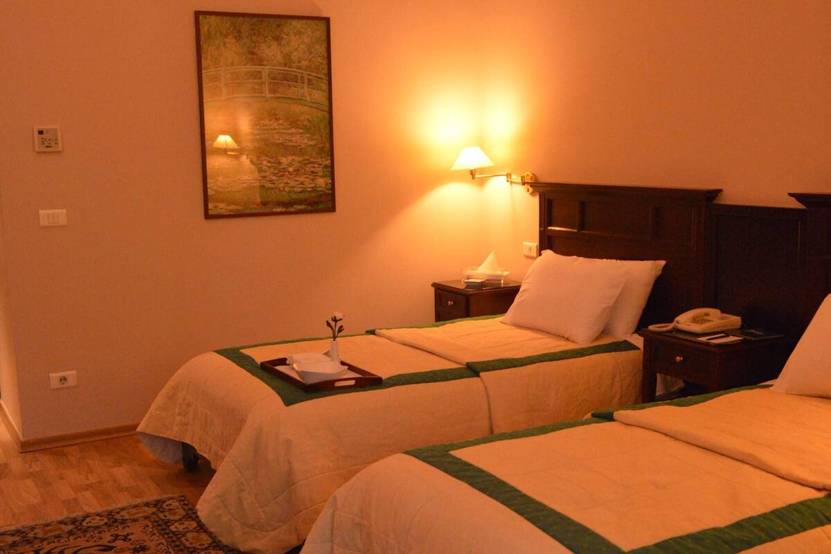 A hotel room with two beds, a nightstand with a lamp, a painting on the wall, and a patterned rug.