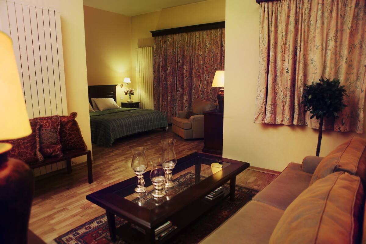A hotel room with a bed, a sofa, a coffee table with glasses, a lamp, and curtains.