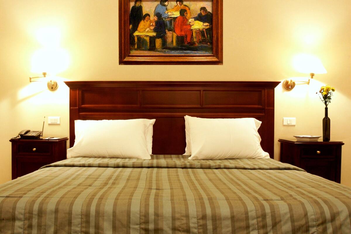 A hotel room with a large bed, two bedside tables, a nightstand, a painting on the wall, and a vase with flowers on the nightstand.