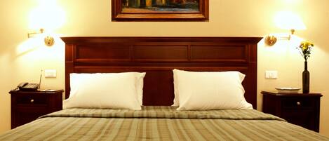 Habitación de hotel con una cama grande, dos mesitas de noche, una mesita auxiliar, un cuadro en la pared y un jarrón con flores sobre la mesita.
