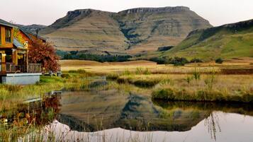 A rustic cabin with a reflective pond, surrounded by rolling hills and a mountain in the background.