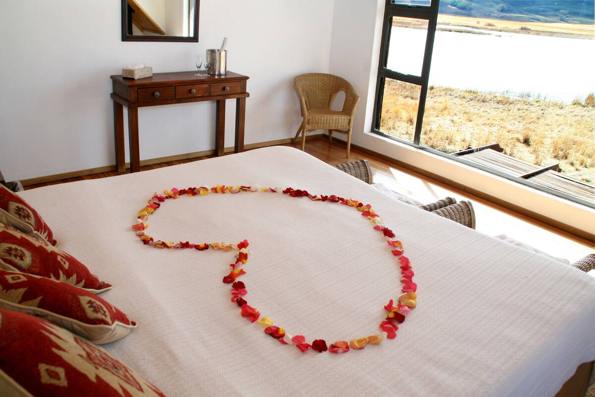 A neatly made bed with a decorative flower arrangement, a wooden nightstand, a wicker chair, and a large window with a view of a field.