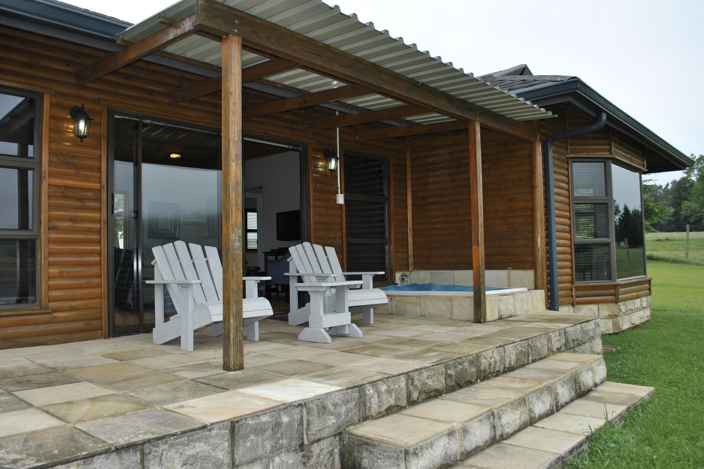 A wooden cabin with a covered patio, two Adirondack chairs, a stone-tiled floor, and a hot tub.