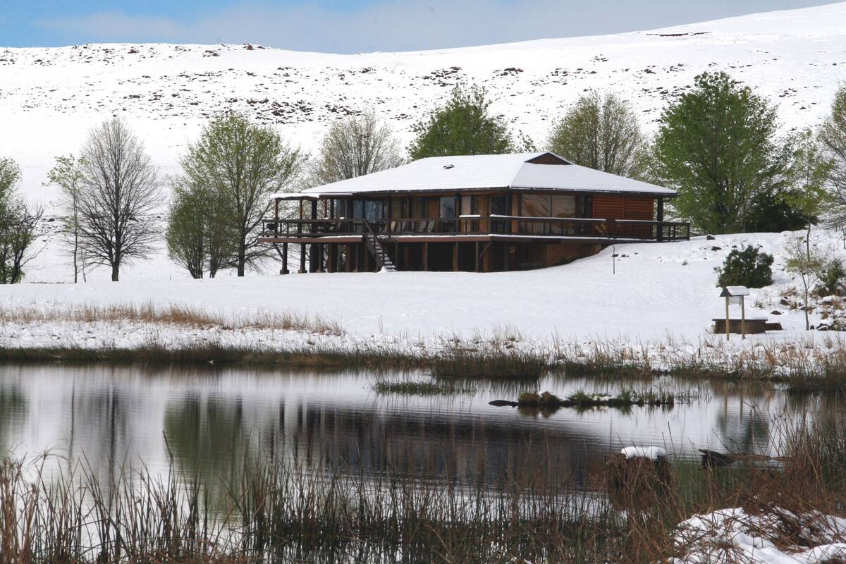 A lodge with a deck overlooking a lake, surrounded by snow-covered hills and sparse trees.