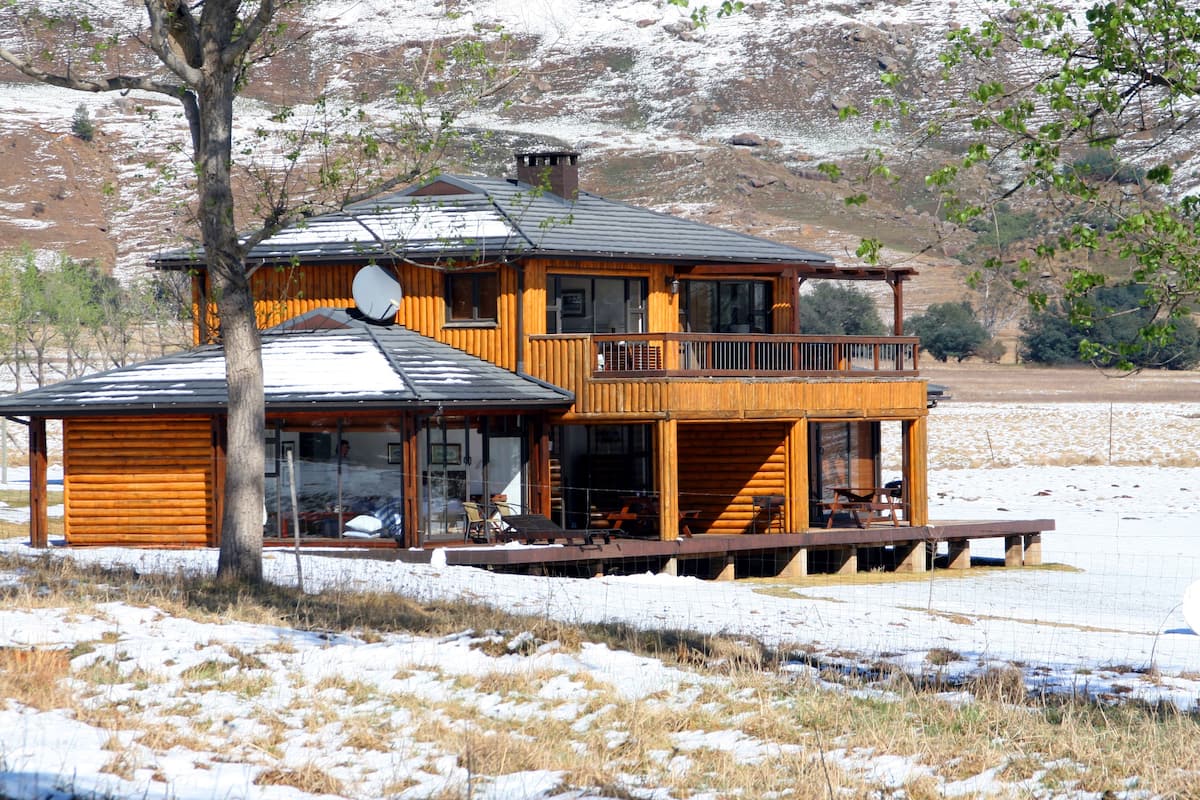 A two-story log cabin with a balcony and a deck, surrounded by snow and trees.