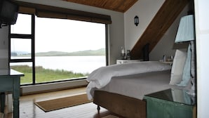 Quarto com uma grande janela com vista para um lago, uma cama, uma mesa de cabeceira e um abajur.