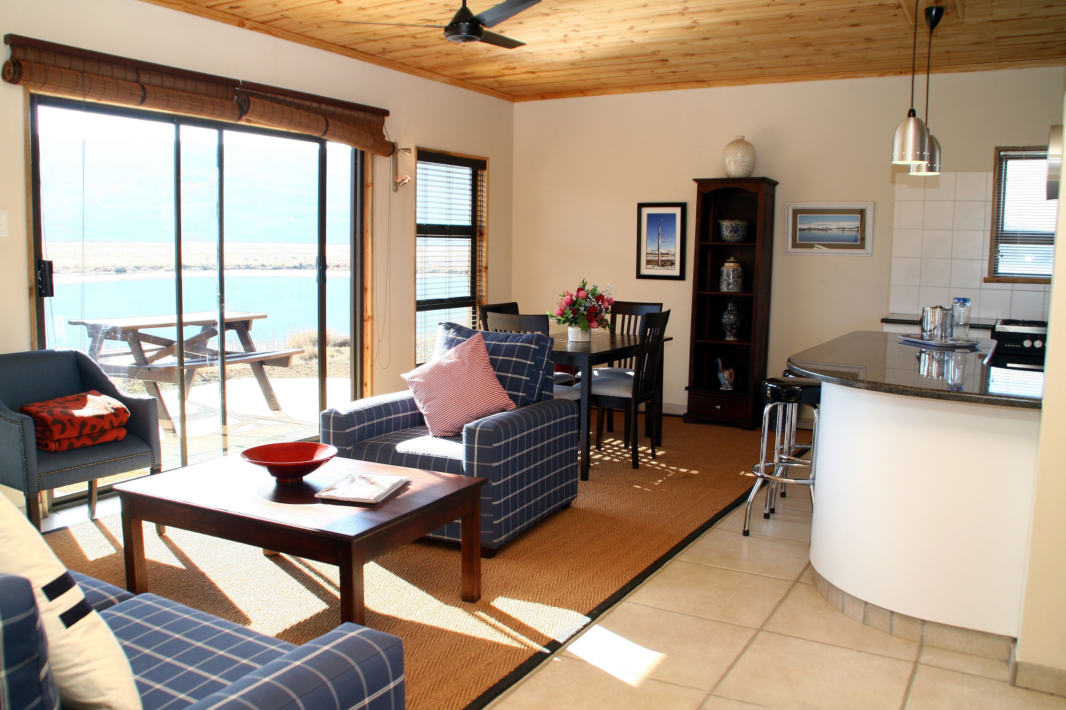 A spacious living area with a sofa, a coffee table, and a dining table. There is a kitchen area with a counter and bar stools. Large windows provide a view of the outdoors.