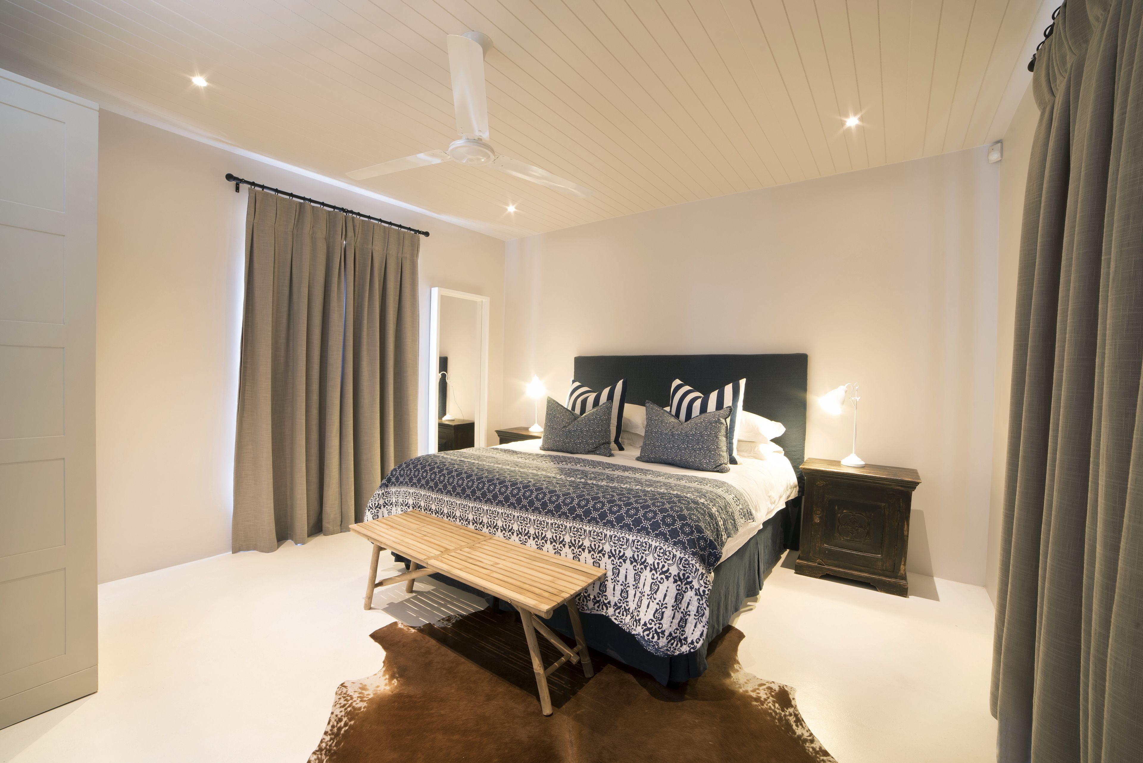 Quarto com cama, mesas de cabeceira, um banquinho e um ventilador de teto.