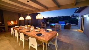 Un comedor al aire libre con una mesa de madera dispuesta para seis personas, sillas blancas y lámparas colgantes.