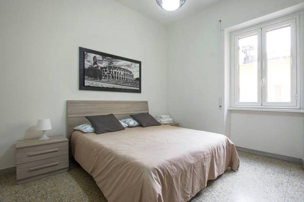 A bedroom with a bed, a nightstand, a lamp, a framed picture on the wall, and a window.