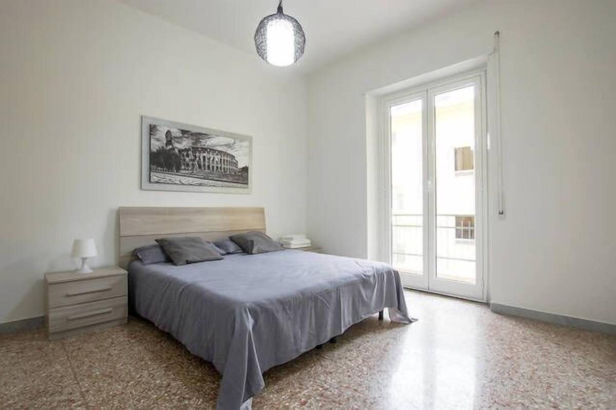A bedroom with a bed, a nightstand, a lamp, a framed picture on the wall, and a large window.