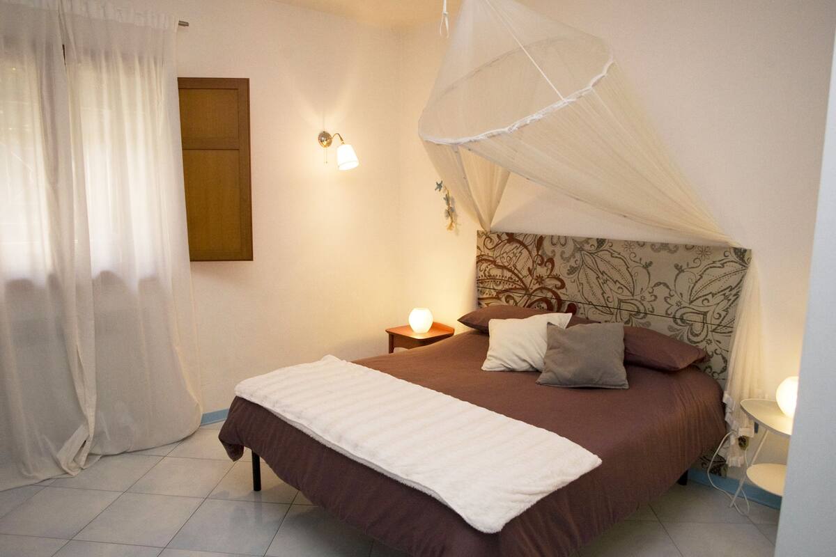 A bedroom with a bed, a nightstand, a wall-mounted lamp, and a window with curtains.