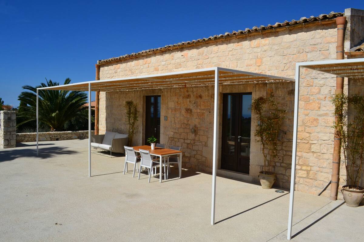 A stone building with a patio area featuring a table and chairs, a covered seating area, and potted plants.