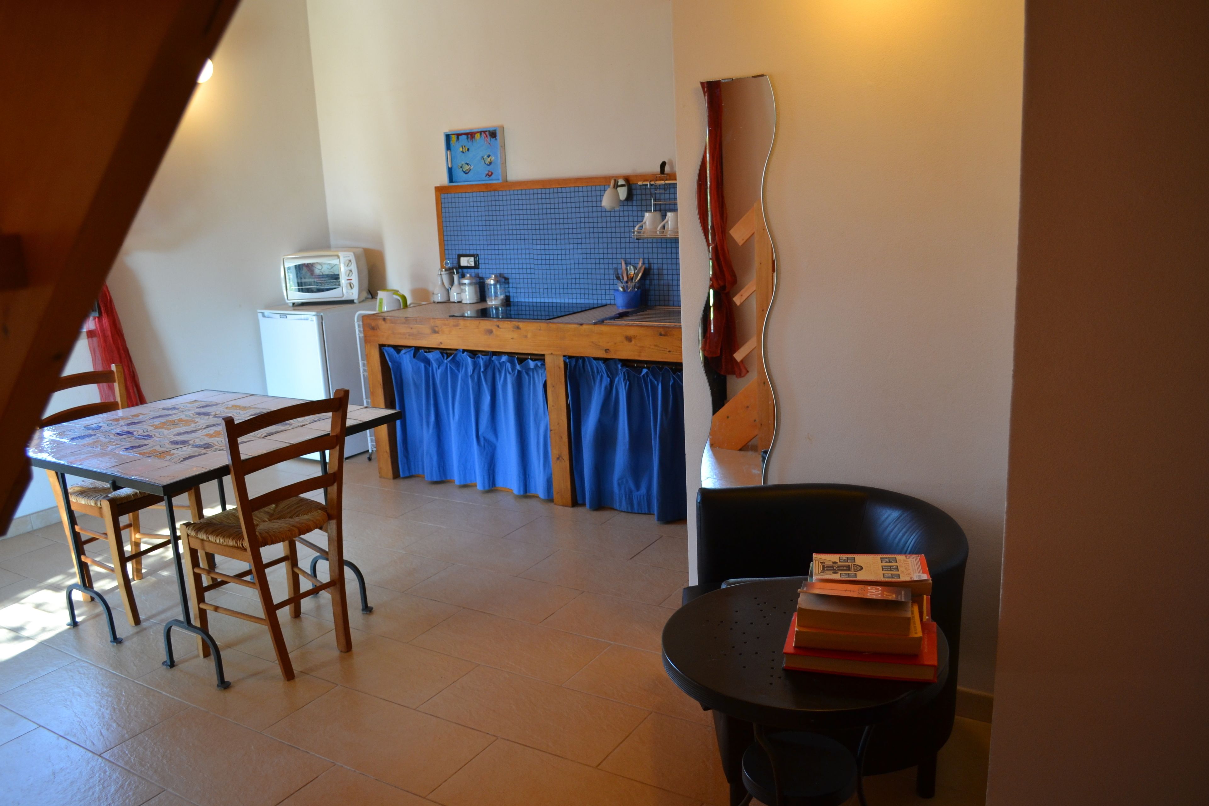 Un coin repas avec une table et des chaises en bois, une cuisine à l’arrière-plan avec des rideaux bleus, et une chaise noire sur laquelle sont posés des livres.