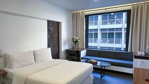 A hotel room with a bed, a small table, a window with a view of a building, and a kitchenette area. - Hamra Urban Gardens (Beirut)