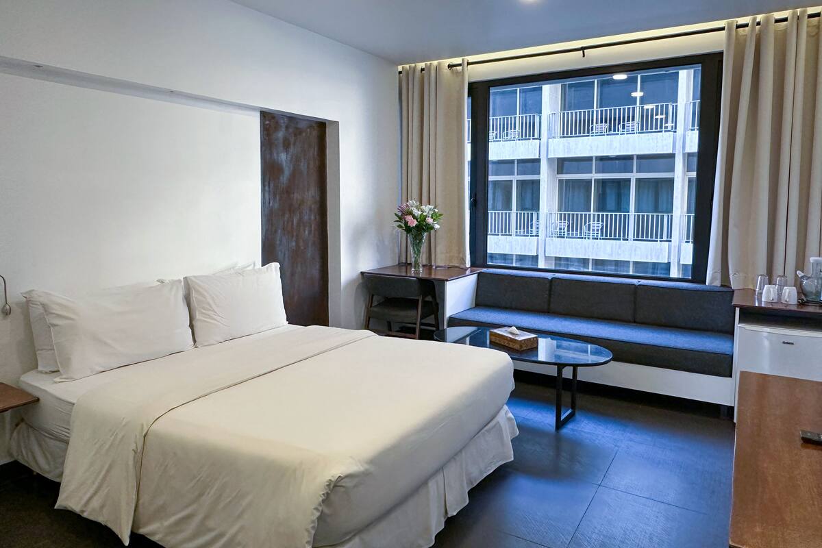 A hotel room with a bed, a small table, a window with a view of a building, and a kitchenette area.