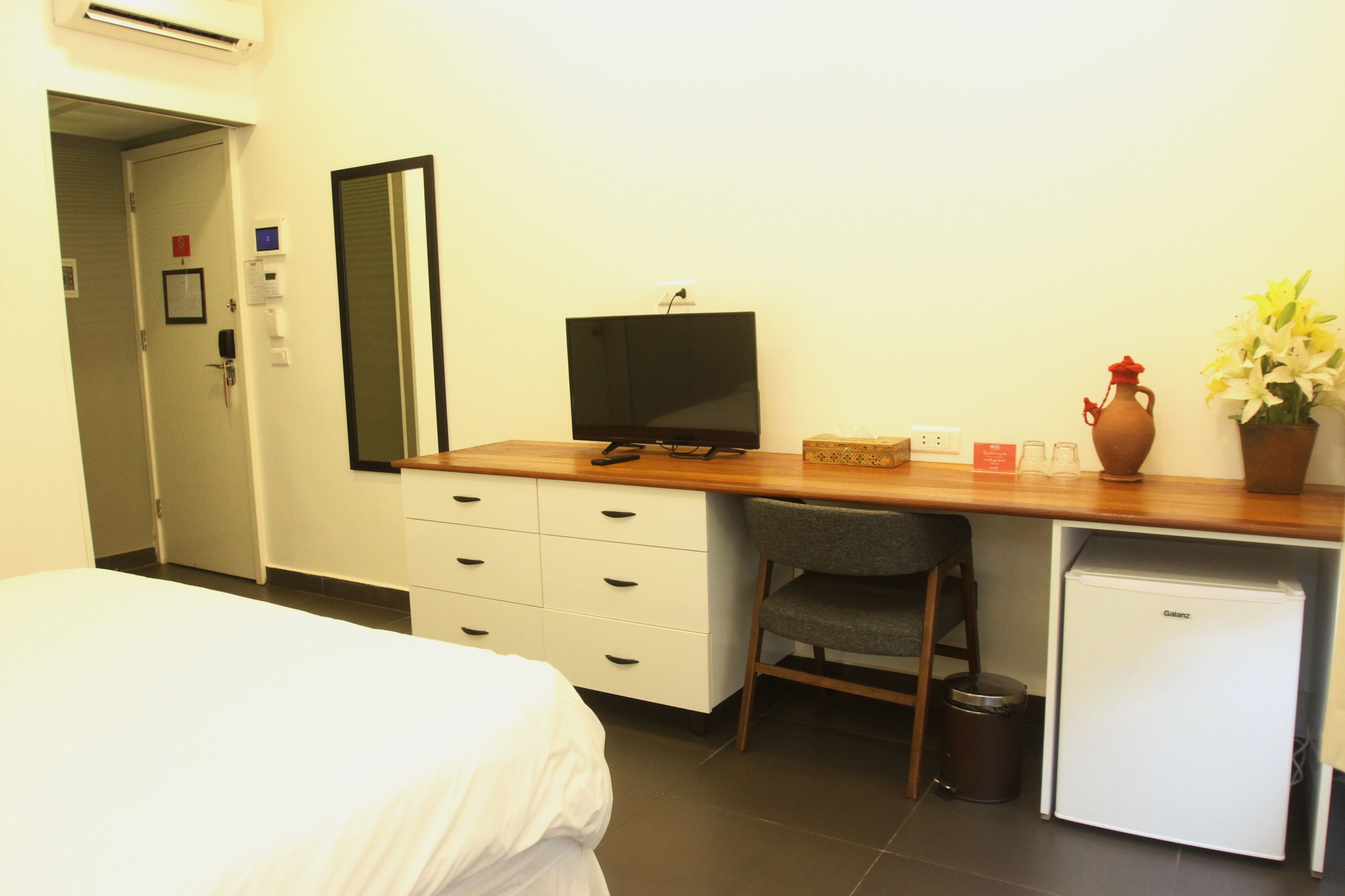 A hotel room with a bed, a desk with a television, a small refrigerator, a chair, and a potted plant.