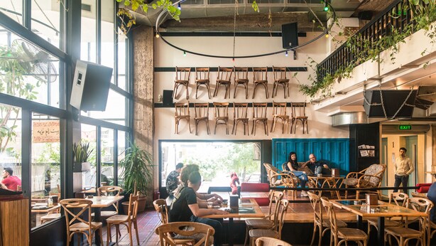 A restaurant with wooden tables and chairs, a staircase, and a view of the street outside. - Hamra Urban Gardens (Beirut)