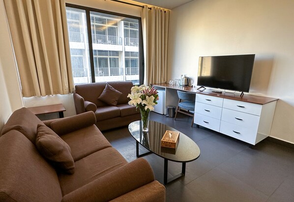 A modern living room with a brown sofa, a glass coffee table, a flat-screen TV, and a dining area with a white cabinet and a small table.