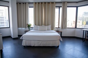 A hotel room with a large bed, two bedside tables, a small table, and a window with curtains.