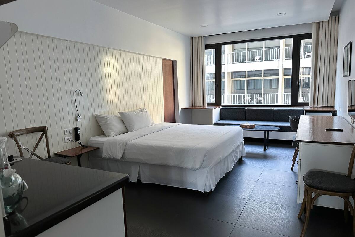 A modern hotel room with a bed, a desk, a chair, a small table, and a window with a view of a building.
