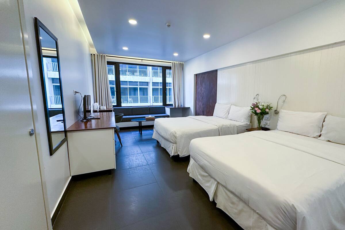 A hotel room with two beds, a desk, a chair, a large window with a view of a building, and a mirror on the wall.