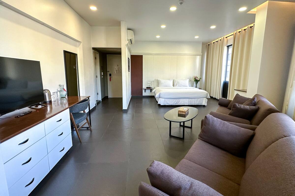 A modern hotel room with a flat-screen TV, a sofa, a coffee table, a bed, and a bathroom visible through an open door.