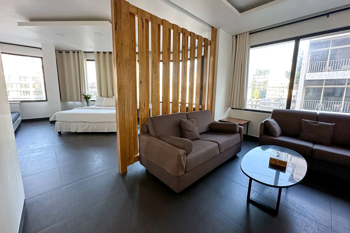 A modern hotel room with a large bed, two sofas, a glass coffee table, and a wooden partition.
