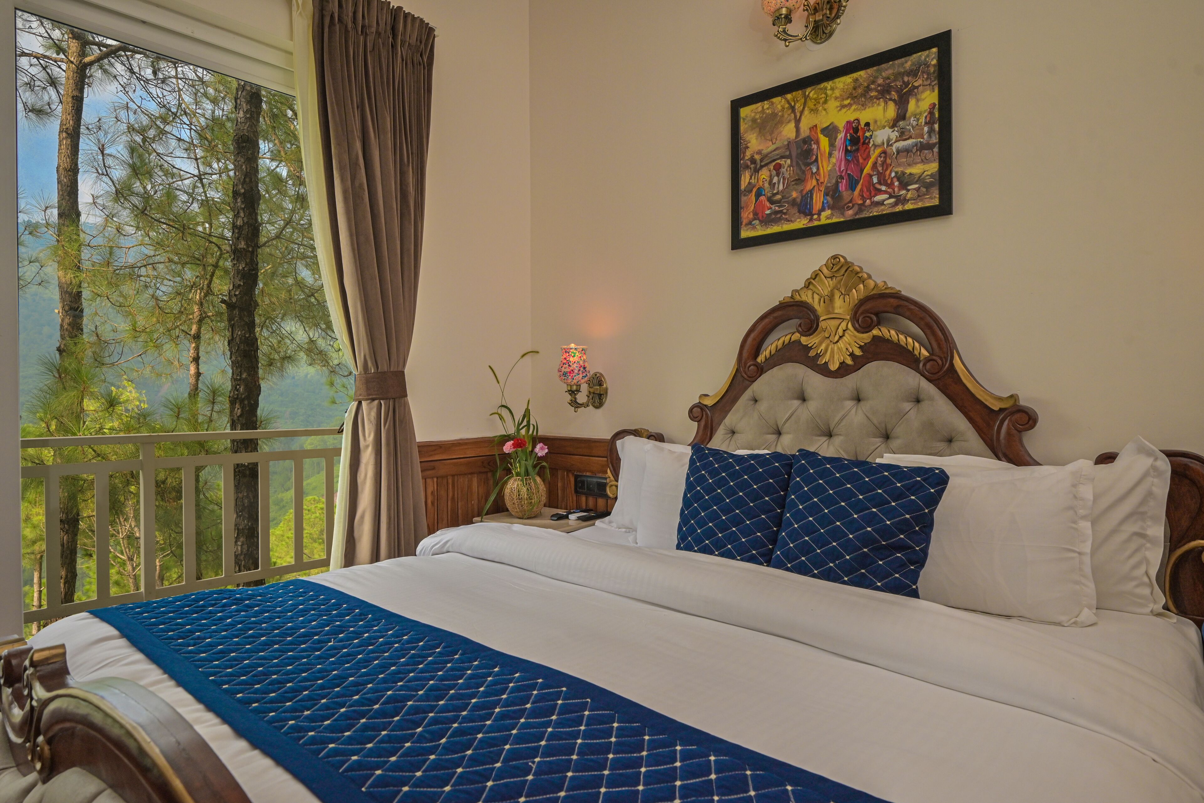 A bedroom with a large bed, a view of trees through a window, a painting on the wall, and a nightstand with a lamp.