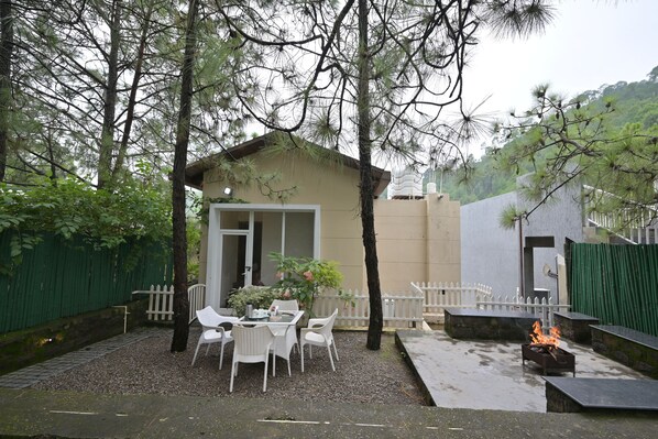 A small outdoor seating area with a table and chairs, a fire pit, and a building with a white fence in the background. - Jungle Lodge Resort (Kasauli)