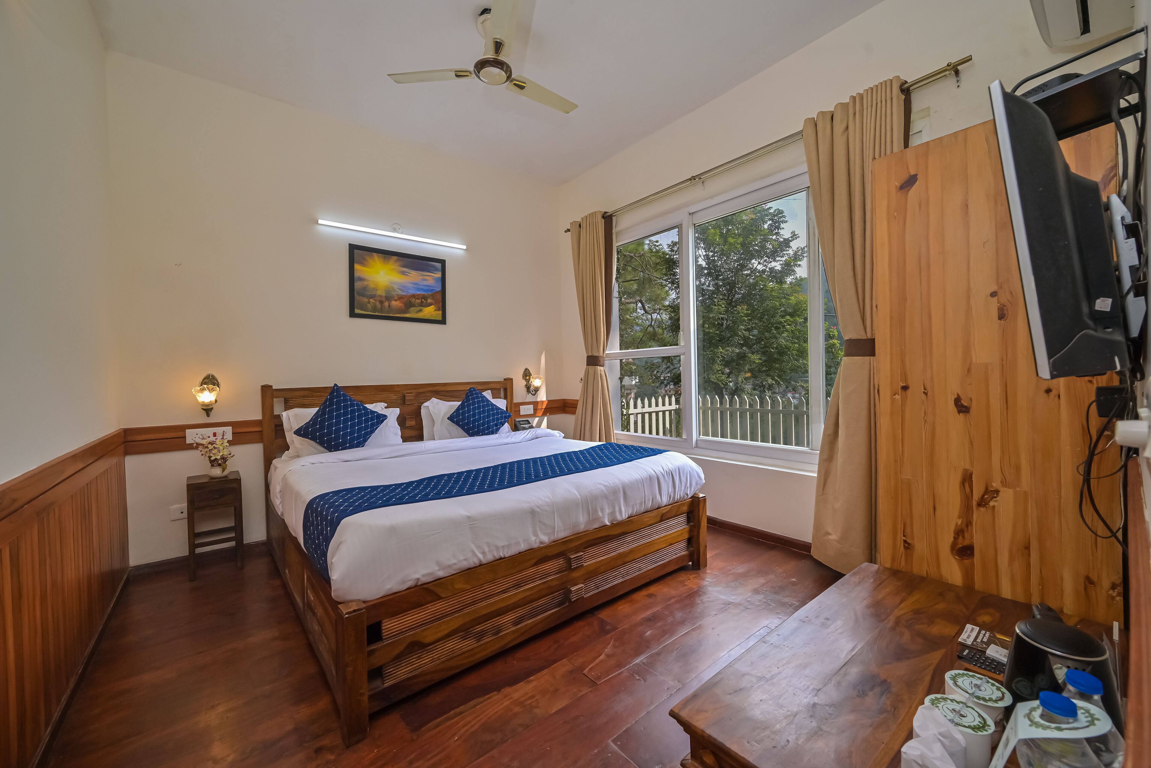 A bedroom with a wooden bed, a nightstand, a ceiling fan, a window with curtains, and a TV mounted on the wall.